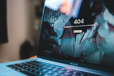 Auf dem Bild ist ein geöffneter MacBook Pro Laptop zu sehen, dessen Bildschirm eine 404-Fehlerseite anzeigt. Die Fehlermeldung lautet "404 - Hmm, the page you were looking for doesn't seem to exist anymore" mit einem Button "Back to Unsplash".  Das Bild hat eine künstlerische, leicht unscharfe Ästhetik mit bläulichen Farbtönen. Man erkennt die Tastatur des MacBook Pro im Vordergrund, während der Bildschirm im Fokus steht und die 404-Fehlermeldung deutlich sichtbar ist. Der Hintergrund ist bewusst unscharf gehalten, was den Fokus auf den Laptop und die Fehlermeldung lenkt.  Es handelt sich um eine stilisierte Darstellung eines typischen Internet-Phänomens - der "Seite nicht gefunden"-Meldung, die jeder Internetnutzer kennt.